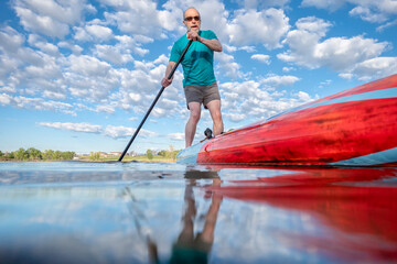  Senior male paddling a stand up paddleboard on a calm lake in Colorado - low angle view from action camera. Recreation, training and fitness concept.