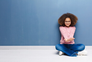 Wall Mural - african american little girl smiling happily with friendly, confident, positive look, offering and showing an object or concept sitting on the floor