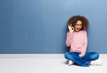 Wall Mural - african american little girl feeling successful and satisfied, smiling with mouth wide open, making okay sign with hand sitting on the floor