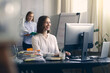 © Martin Villadsen - Attractive and professional young female employee working on the computer at the office while her coworker and coleague is walking in the background at the office.