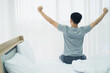 © linghaa - Young Asian man wake up from bed in white bed room which felling happy. Corona virus, COVID-19 Pandemic new normal life at home Concept.