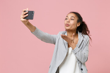 Wall Mural - Pretty african american business woman in grey suit, white shirt isolated on pink background. Achievement career wealth business concept. Doing selfie shot on mobile phone, blowing sending air kiss.