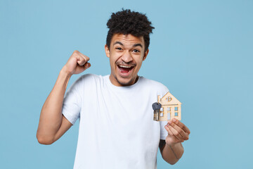 Wall Mural - Happy young african american guy in casual white t-shirt posing isolated on blue background in studio. People lifestyle concept. Mock up copy space. Hold house bunch of keys, doing winner gesture.