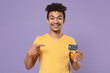 © ViDi Studio - Smiling young african american guy in casual yellow t-shirt posing isolated on pastel violet background studio. People lifestyle concept. Mock up copy space. Pointing index finger on credit bank card.