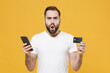 © ViDi Studio - Shocked young bearded man guy in white casual t-shirt posing isolated on yellow wall background studio portrait. People lifestyle concept. Mock up copy space. Hold credit bank card using mobile phone.