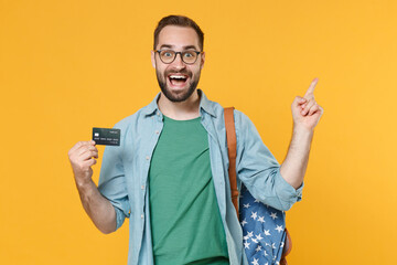 Excited young man student in casual clothes glasses with backpack isolated on yellow background. Education in high school university college concept. Hold credit bank card pointing index finger up.