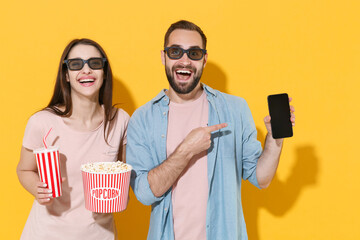 Wall Mural - Excited couple friends guy girl in 3d glasses isolated on yellow background. People in cinema concept. Watching movie film, hold bucket popcorn, cup soda point on mobile phone with blank empty screen.