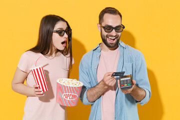 Wall Mural - Shocked couple friends guy girl in 3d glasses isolated on yellow background. People in cinema concept. Watching movie film, hold popcorn cup soda bank terminal to process acquire credit card payments.