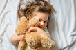 © kaloriya - Smiling toddler girl holding teddy bear and lying in bed on white sheets at home. Happy child. Top view.