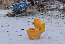 Tiny Yellow Toy Toilet On The Ground In The Snow At A Garbage Dump.  Piles Of Garbage In The Background.  Sad Discarded Toy, But Weird!