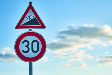 A red and white traffic sign, which warns of a twelve percent gradient and a traffic sign below it with a speed limit of 30 km/h. A blue sky with white clouds is in the background.