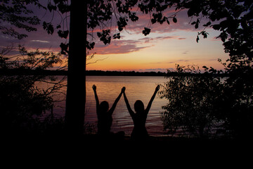 silhouette of two happy girls sitting together holding hands with raised arms looking towards a beautiful purple and orange sunset from the beach to the shore of on a large lake next to a tree