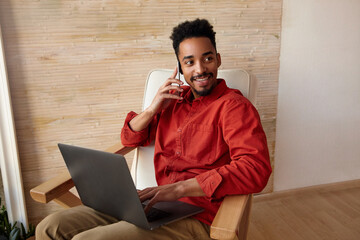 Wall Mural - Cheerful young lovely short haired bearded male with dark skin smiling widely aside while making call, working remotely from home, isolated over beige background