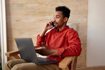 Wall Mural - Indoor photo of young short haired bearded guy with dark skin having phone conversation while sitting in front of window in chair, working out of office