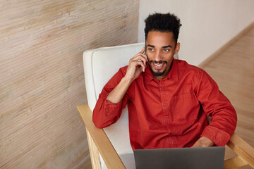 Wall Mural - Joyful young attractive brunette bearded dark skinned male smiling happily while having nice phone talk, sitting in chair with laptop over beige interior