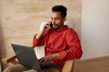 Wall Mural - Indoor photo of young dark skinned businessman with short curly hair frowning his eyebrows during phone talk and looking attentively on screen of his laptop, isolated over home interior