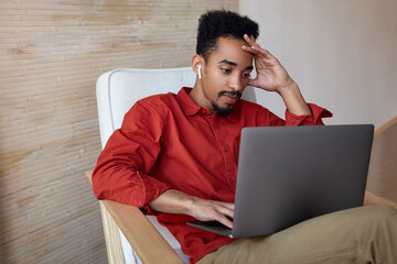 Wall Mural - Concentrated young bearded dark skinned man with short haircut keeping raised hand on his head while checking email box on his laptop, isolated over home interior