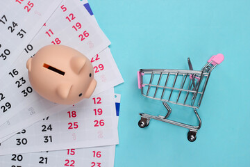 Wall Mural - Seasonal discounts. Piggy bank with monthly calendar, supermarket trolley on a blue background. Top view