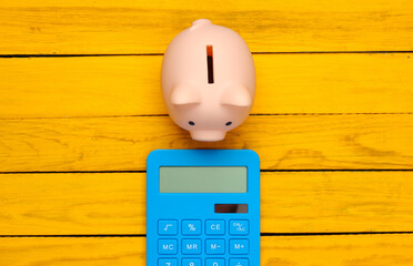 Piggy bank and blue calculator on a yellow wooden background. Top view