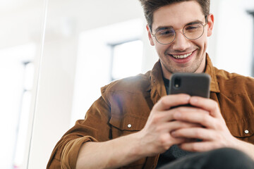 Image of joyful young man smiling and using cellphone while working