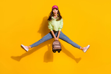 Wall Mural - Full size photo of funky energetic attractive lady student good mood hold backpack jump high up wear green cropped sweatshirt jeans shoes cap isolated vivid bright yellow color background