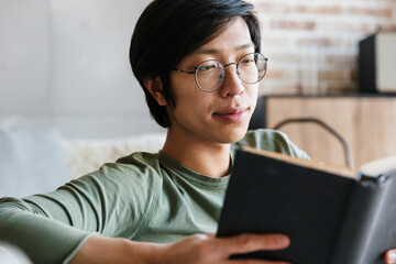 Canvas Print - Image of young asian man wearing eyeglasses reading book in apartment