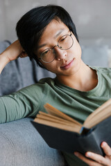 Wall Mural - Image of young asian man wearing eyeglasses reading book in apartment