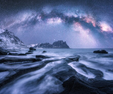 Milky Way Over The Snow Covered Mountains And Rocky Beach In Winter At Night In Lofoten Islands, Norway. Landscape With Purple Starry Sky, Water, Stones, Snowy Rocks, Bright Milky Way. Beautiful Space