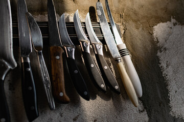 A set of metal knives of different sizes and shapes, hanging on a horizontal magnet holder. On a gray concrete wall made of block and cement