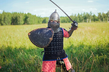 A medieval Central Asian warrior, a nomad in 14th-century armor, stands in a defensive or attacking position with a shield and a saber in his hands in a field against a forest.