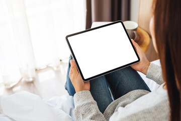 Top view mockup image of a woman holding black tablet pc with blank desktop white screen while sitting on a cozy white bed at home