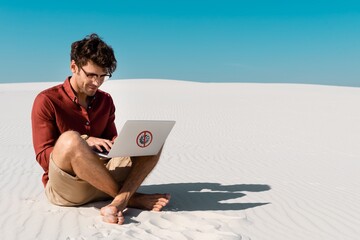 Wall Mural - young freelancer on sandy beach using laptop with stop virus sign against clear blue sky