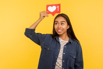 Social media button. Portrait of thoughtful girl in denim shirt holding heart icon on head, follower notification symbol, dreaming of many Likes and internet popularity. indoor studio shot isolated