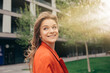 © Anton - Cheerful positive happy young woman look up and back with smile. Walking alone outside on street during sunshine. Stylish trendy girl wears orange jacket. Summertime or spring season.
