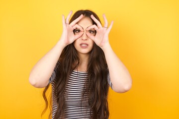 Wall Mural - Playful excited teenage female with dark long hair showing Ok gestures with both hands, pretending to wear spectacles, astonished to see something amazing. Emotions, body language and gesturesï¿½