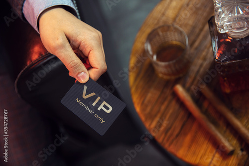 Man holds VIP member card. View from the top on the gentleman\'s hand that holds exclusive VIP membership card next to the wooden table with whisky in carafe and glass with cigars.