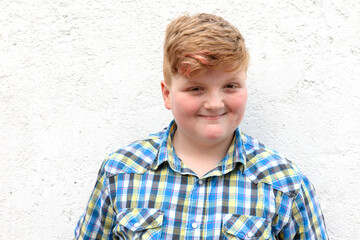 Plump red-haired boy is smiling. A boy in a plaid shirt stands on a wall background and smiles.