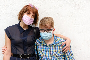 Sister wearing trendy glasses and little brother with glasses posing together looking at the camera.Young red-haired woman and boy wearing a surgical mask stand against a wall background.