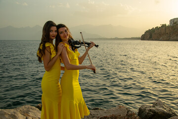 Wall Mural - Two beautiful girls violinists in yellow concert dresses are playing electric violins with sunset, mountains and Mediterranean sea on the background. Antalya Old Town Kaleici, Turkey.  Stock image.