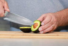 Male Hands About To Remove The Seed Of A Freshly Cut Avocado With A Kitchen Knife On A Wooden Cutting Board.