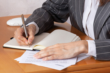 Close up hands of business woman wearing gray striped suit, and writing with expensive pen in notebook lying on wooden table. Paper documents, white computer mouse, light background wall. Indoors.