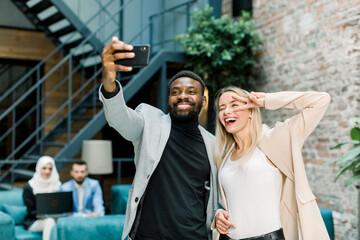 Wall Mural - Happy business people, working in office. Smiling Caucasian lady and African man making joint selfie photo on smartphone, posing, laughing and showing dofferent gestures