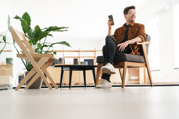 Wall Mural - Image of joyful young man smiling and using cellphone while working