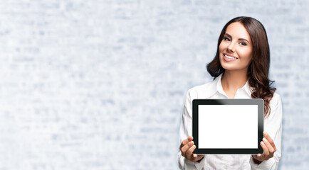 Canvas Print - Beautiful brunette confident businesswoman showing blank no-name tablet pc monitor, with copy space for some text. White brick wall background.