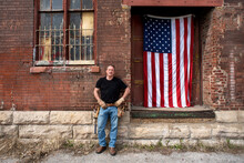 Male Blue Collar Worker In Tee Shirt Standing Next To Old Brick Factory Loading Dock Wearing Tool Belt American Flag Hanging On Door