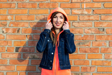 Happy young woman in denim jacket, puts a hood on her head on a background of a red brick wall