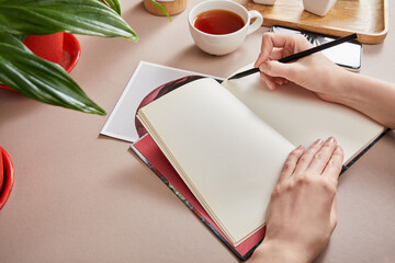 Poster - cropped view of woman writing in planner near green plant, cup of tea, smartphone on beige surface