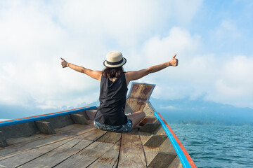 Back view of woman wearing hat sitting on the head of  boat, 
extend the arms out to sides with thumbs up and enjoying holiday on the boat , traveling concept. 