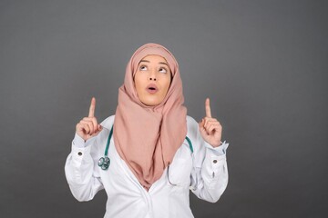 Canvas Print - Portrait of beautiful doctor woman over isolated background being amazed and surprised looking and pointing up with fingers showing something strange.