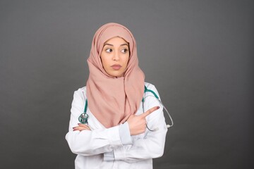 Wall Mural - Excited surprised muslim girl wearing hijab and medical uniform points happily aside on copy space of gray background for your advertising information or promotional text.